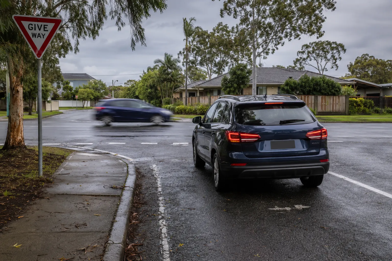 A car waiting at a give way line at a wet suburban intersection with traffic passing across in front of it